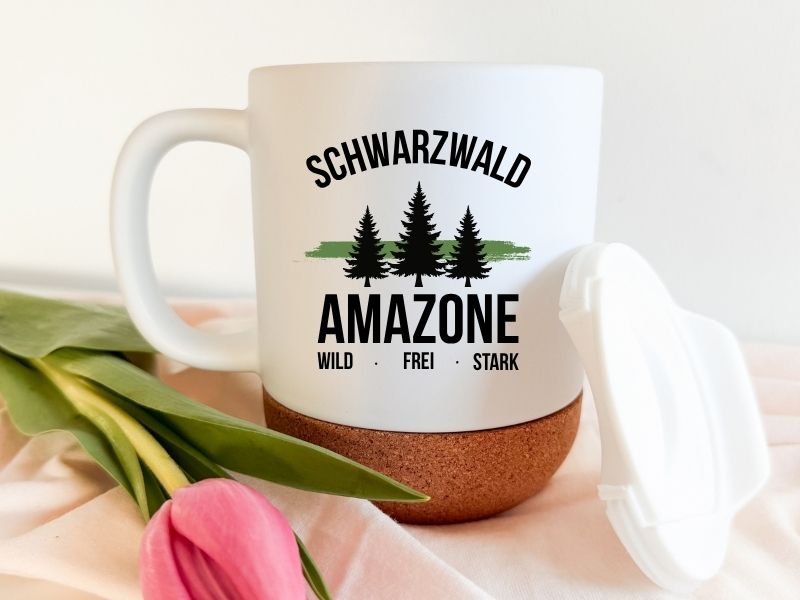 Weiße Keramiktasse mit Korkboden und Deckel, Aufdruck 'Schwarzwald Amazone - Wild Frei Stark' mit Tannenbaum-Motiv, neben einer rosa Tulpe